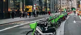 A row of green rental bikes is lined up along a city street, with modern glass buildings and people walking on the sidewalk. The scene appears to be in a busy urban area with high-rise offices.