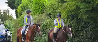 Two people, wearing helmets and reflective vests, are riding brown horses along a quiet, paved road bordered by lush green hedges. In the background, a parked blue car is partially visible. The riders appear to be relaxed and enjoying their leisurely ride.