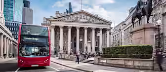 A red double-decker bus with the destination "London Bridge" displayed on its sign passes in front of the Royal Exchange in London. Modern office buildings and a statue of a man on horseback are visible in the background under a partly cloudy sky.