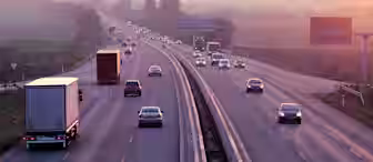 A wide highway with multiple lanes in each direction is pictured during sunset. Several cars and trucks are driving on both sides of the road, with a scenic countryside and trees in the background. The sky displays beautiful hues of pink, purple, and orange.
