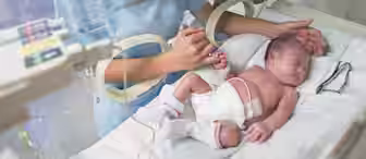 A nurse gently holds the hand of a newborn baby inside an incubator in a hospital neonatal unit. The baby is lying on a white pillow, wearing a diaper and small socks, connected to medical equipment.