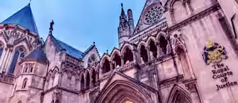 Image of the exterior of The Royal Courts of Justice in London, featuring Gothic architecture with intricate arches, pointed spires, and detailed carvings. The building's name is visibly inscribed on the wall along with a crest, illuminated by warm lighting from within.