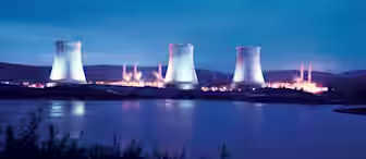 A nuclear power plant with four cooling towers illuminated at dusk. The plant is situated near a body of water with lush greenery in the foreground and mountains in the background, all under a twilight sky.