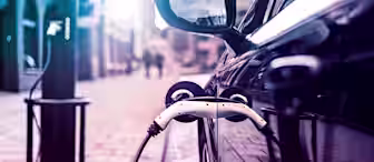 A close-up shot of an electric vehicle being charged at a charging station on a city street. The car's side mirror and charging port are prominently in view, with the plug connected. The background is slightly blurred, showing buildings and streetlights.