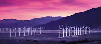 A field of wind turbines stretches across a desert landscape with mountains in the background, under a vibrant pink and purple sunset sky. The turbines are aligned in rows, and the desert floor is sparsely covered with vegetation.