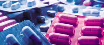 Several packs of pills in various shapes and colors are scattered on a surface. The medication is contained in blister packs, with some capsules being half white and half blue, others in different color combinations. The image uses blue and pink lighting.