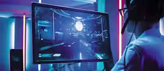 A person wearing glasses and headphones is sitting at a desk, playing a futuristic video game on a large monitor. The room is illuminated with blue and pink neon lights, and speakers are placed on both sides of the monitor.