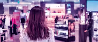 A person with long hair, seen from behind, stands in a brightly lit, modern store. The store is filled with various displays of clothing and cosmetics, with shelves and racks visible in the background. The atmosphere is vibrant and bustling.