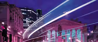 A nighttime cityscape featuring the Royal Exchange in London, illuminated with pink and blue lights. Light trails from passing vehicles create dynamic streaks in the foreground. Surrounding buildings are also visible under a deep blue and purple sky.