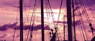 Silhouettes of ship masts and rigging against a vibrant, purple-hued sunset sky with scattered clouds. The dark outlines create a dramatic contrast with the soft and colorful background.