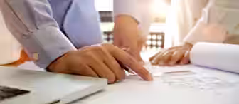 Two people wearing light blue shirts are reviewing architectural blueprints on a table in a brightly lit room. One person is pointing at the blueprints with a pen, while the other rests their hands on the table. A laptop is visible to one side.