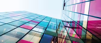 A low-angle view of a modern building with colorful, reflective glass panels. The vibrant sections of red, pink, blue, and yellow stand out against the clear sky, creating a visually striking and geometric pattern.