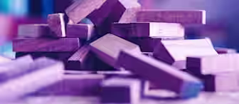 A close-up view of a pile of scattered rectangular wooden blocks on a surface. The blocks are of various lengths and widths and are stacked haphazardly, with a soft purple hue illuminating the scene. The background is blurry, emphasizing the foreground focus.