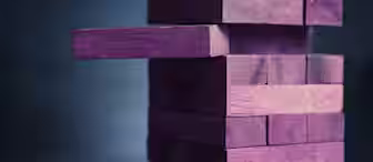 A close-up view of a tower made of purple wooden blocks in the style of the game Jenga. One block is partially pulled out from the middle of the stack, creating an imbalance. The background is slightly blurred and dark.