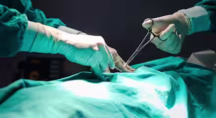 Two surgeons in green surgical gowns and gloves perform a procedure on a patient, using surgical instruments under focused lighting.