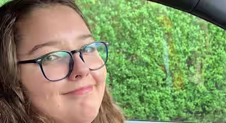 A person with long hair wearing glasses, a dark jacket, and a white shirt sits in the driver's seat of a car, smiling at the camera. Green foliage is visible through the driver's side window.