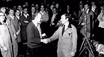 A black-and-white photo of a group of men in a workshop. Two men in the center are shaking hands, surrounded by others who are smiling and watching. The setting appears casual, with some men in overalls and work clothes.