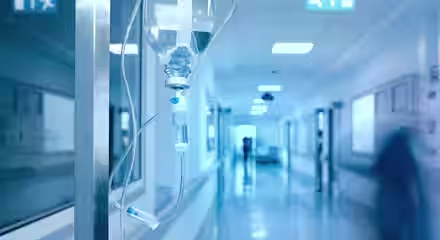 A close-up of an intravenous (IV) drip in a hospital corridor. The IV drip hangs from a metal pole and is partially filled with clear fluid. The corridor is modern, well-lit, and slightly blurred, with people in motion in the background.