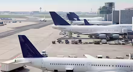 Several large commercial airplanes are parked at the gates of a busy airport terminal. Various ground service vehicles are attending to the planes, and the airport runway can be seen in the background under a clear blue sky.