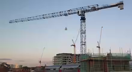 A construction site with three cranes, including a large blue and white tower crane, working on several buildings. The structures are surrounded by scaffolding and green netting. The background shows a clear sky during sunset, with buildings in the distance.