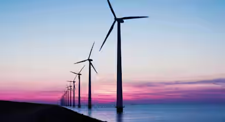 A row of wind turbines stands tall against a vibrant sunset backdrop over a body of water. The sky is painted with hues of pink, purple, and blue as the day transitions into evening. The turbines are aligned in perspective, stretching into the distance.