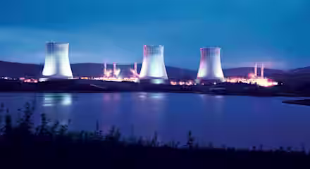 A nuclear power plant with four cooling towers illuminated at dusk. The plant is situated near a body of water with lush greenery in the foreground and mountains in the background, all under a twilight sky.