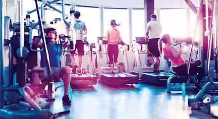A group of people exercising in a gym. Two men and two women are using treadmills in the background. In the foreground, one man is using a weight machine, and one woman is performing pull-ups. The gym is brightly lit with large windows.