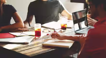 A group of four people seated around a wooden table engaged in a discussion. The table has a laptop, notebooks, glasses of iced tea, and documents scattered across it. Sunlight is streaming through a window, creating a bright atmosphere.