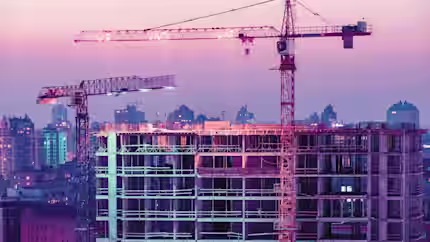 Construction site featuring two large cranes and an unfinished multi-story building under a colorful dusk sky. The background displays a cityscape with various buildings illuminated by the transitioning evening light, creating a blend of urban progress and twilight hues.