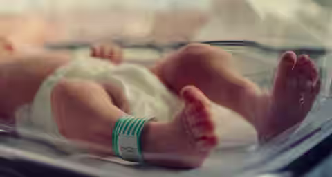 A newborn baby lies in an incubator, wearing a diaper and an identification wristband. The baby's feet are in focus, slightly elevated, and toes are clearly visible. The background is soft and out of focus, emphasizing the fragile and delicate state of the infant.