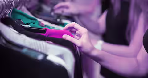 Close-up of people browsing through clothes on a rack. One hand is prominently seen touching a pink garment, while other blurred figures in the background also look through clothing items. The scene appears to be in a store or a market with a purple-tinted lighting.