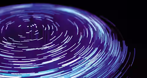 A circular, abstract light pattern with bright blue and purple streaks radiating outward on a dark background, resembling long-exposure photography of spinning lights. The streaks create a sense of motion and depth, forming a visually dynamic composition.