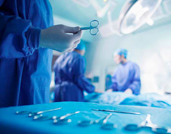 A surgeon in blue scrubs and gloves holds a surgical tool, possibly a pair of forceps, while standing next to an operating table. In the background, two other surgeons in blue scrubs and masks work on a patient under bright operating room lights. Various surgical instruments are laid out on a table in the foreground.