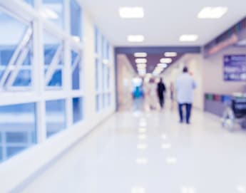 A blurred image of a hospital corridor with bright lighting. People are walking in the distance, and a wheelchair is positioned near a counter on the right side. Large windows on the left let in natural light. Tables with information are on the right side of the hallway.