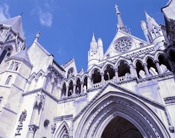 Gothic-style facade of the Royal Courts of Justice in London, featuring ornate detailing, pointed arches, and a large rose window under a clear blue sky. The architecture includes a mix of spires and turrets.