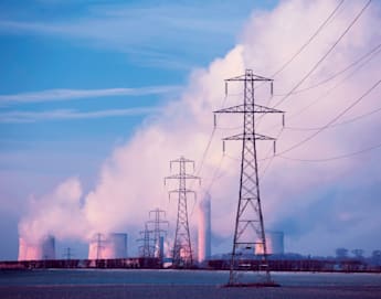 Power lines stretch across a field towards a power plant with cooling towers emitting white steam against a blue sky. The scene is illuminated by the soft pink and purple hues of either sunrise or sunset, contrasting with the industrial structures.