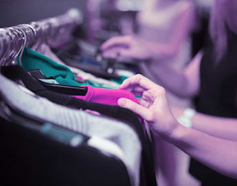 Close-up of people browsing through clothes on a rack. One hand is prominently seen touching a pink garment, while other blurred figures in the background also look through clothing items. The scene appears to be in a store or a market with a purple-tinted lighting.