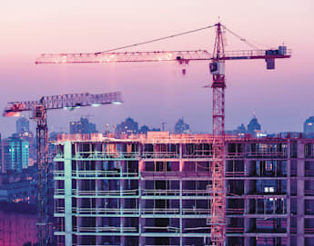 Construction site featuring two large cranes and an unfinished multi-story building under a colorful dusk sky. The background displays a cityscape with various buildings illuminated by the transitioning evening light, creating a blend of urban progress and twilight hues.