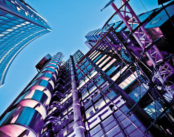 A low-angle view of modern high-rise buildings against a clear blue sky. The structures feature a mix of glass, steel, and vibrant purple lighting, showcasing architectural details with a futuristic appearance.