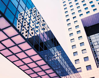 A modern architectural structure featuring two tall white buildings with numerous windows, connected by a glass bridge. The facade of one building reflects the other. The image is taken from a low angle with a clear blue sky in the background.