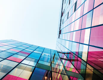 A low-angle view of a modern building with colorful, reflective glass panels. The vibrant sections of red, pink, blue, and yellow stand out against the clear sky, creating a visually striking and geometric pattern.