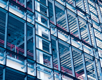 Close-up of a modern building's glass facade, revealing its structural framework. The geometric design features a grid of metal beams and glass panels, with red railings and metal conduits visible within the structure. The image has a blue tint.