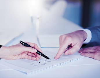 Two people, one in a dark suit and the other in a light suit, are seated at a table. One person is holding a pen and pointing to a document on the table, while the other person is pointing at the same document. The scene suggests a discussion or review of the document.