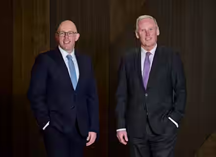 Two men in suits stand smiling against a dark wooden background. The man on the left wears glasses and a blue tie, while the man on the right wears a purple tie. Both have their hands in their pants pockets.