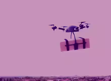 A drone carrying a package flies over a cityscape with a pink sky. The horizon features distant buildings and trees.