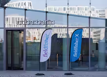 The entrance to Cube Berlin with reflective glass walls, showcasing two blue and white flags bearing the "fieldfisher" logo. The modern building architecture is visible in the background.
