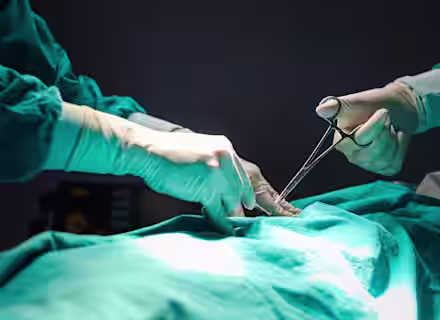 Two surgeons in green surgical gowns and gloves perform a procedure on a patient, using surgical instruments under focused lighting.