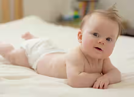A baby with light skin and blond hair lies on its stomach on a bed, propped up on its forearms. The baby is wearing only a diaper and looks directly at the camera, with a hint of a smile. The background is softly blurred, showcasing a cozy indoor setting.