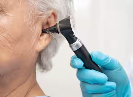 A doctor wearing blue gloves uses an otoscope to examine the ear of an older adult with gray hair during a medical checkup.