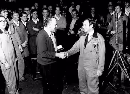 A black-and-white photo of a group of men in a workshop. Two men in the center are shaking hands, surrounded by others who are smiling and watching. The setting appears casual, with some men in overalls and work clothes.